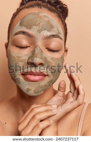 Doing beauty procedure concept. Studio vertical close up of young relaxed pretty cheerful European woman with closed eyes applying clay face mask standing isolated on beige background in undershirt