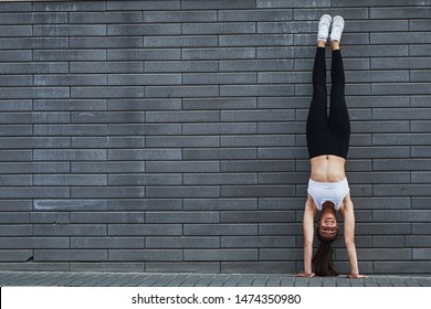 Doing handstand. Young sportive brunette with slim body shape against brick wall in the city at daytime.