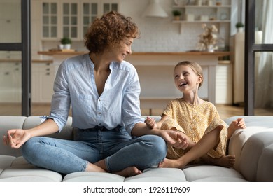 Doing like mommy. Laughing young mother little school age daughter having fun sit cross legged on couch at modern studio flat do yoga together. Funny small kid girl imitate mom in meditation practice