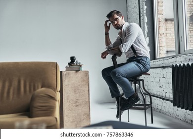 Doing some thinking. Handsome young man looking at camera and touching his head with hand while sitting on stool