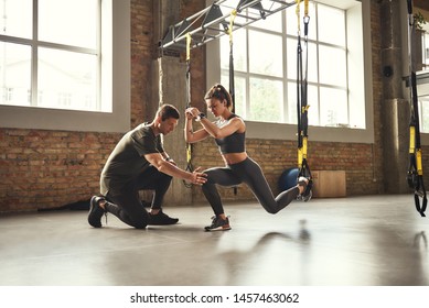 Doing squat exercise. Confident young personal trainer is showing slim athletic woman how to do squats with Trx fitness straps while training at gym.