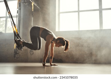 Doing TRX exercises. Young athletic woman in sports clothing training legs with trx fitness straps in the gym. - Powered by Shutterstock