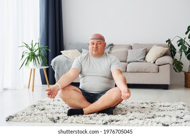 Doing yoga exercises. Funny overweight man in casual clothes is indoors at home.