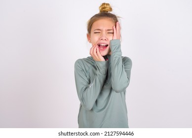 Doleful Desperate Crying Little Caucasian Kid Girl With Hair Bun Wearing Technical Shirt Over White Background , Looks Stressfully, Frowns Face, Feels Lonely And Anxious