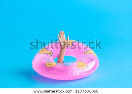 Doll arm with inflatable pool float emerging from blue paper background in form of sea. Drowning minimal creative abstract concept.