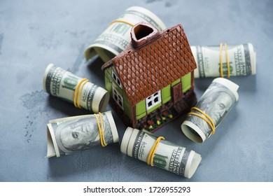 Doll House On A Grey Concrete Background Surrounded By Rolls Of American Dollar Bills, Selective Focus, Studio Shot