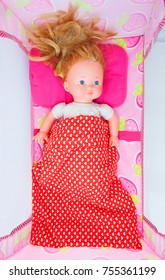 Doll Laying In The Baby Cot. Overhead Shot Of The Doll With Red Hair Laying On Pink Pillow And Covered With Red Baby Duvet. Small Girlish Toy With No Clothes, Blue Eyes Laying In The Pink Baby Bed.