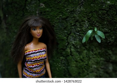 A doll standing in front of a tree trunk with green leaves 