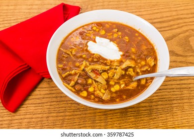 Dollop Of Sour Cream In Spicy Bowl Of Chicken Tortilla Soup On Old Oak Table.  Over Head View With Red Napkin.