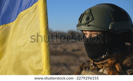 Dolly shot of girl in military helmet and balaclava holds a waving national flag. Female soldier of ukrainian army lifted blue-yellow banner at countryside. Invasion of territory Ukraine. Close up.