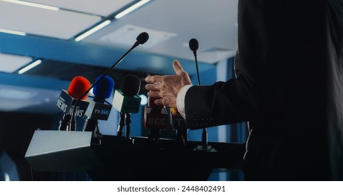 Dolly shot of male organization representative pronouncing speech from the tribune during press campaign in the conference hall. Politician answers questions, gives interview for television or media.