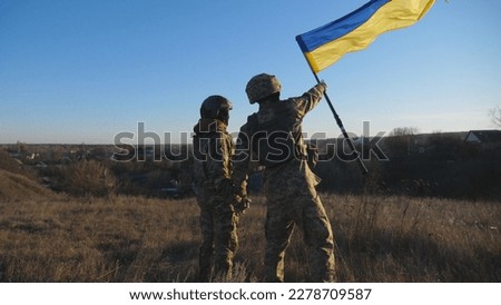 Dolly shot of young woman and man in military uniform waving flag of Ukraine against sunset. Female and male soldier of ukrainian army lifted blue-yellow banner holding hands of each other at field.