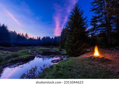 In the Dolly Sods Wilderness in West Virginia, a campfire burns next to the Left Fork of Red Creek where the Blackbird Knob Trail crosses it. Pink clouds reflect in the water at sunset. - Shutterstock ID 2311044623