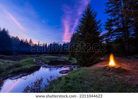 In the Dolly Sods Wilderness in West Virginia, a campfire burns next to the Left Fork of Red Creek where the Blackbird Knob Trail crosses it. Pink clouds reflect in the water at sunset.