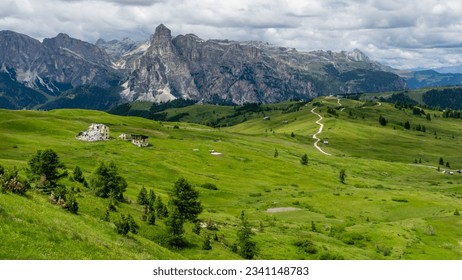 Dolomite mountain range landscape scenery in summer with grassy plains and a ruins structure, some trees, and a hiking trail before craggy peaks during cloudy weather in italy.