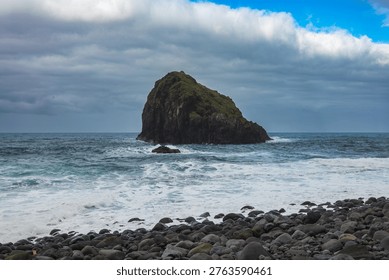 A dome sea stack rises from the Atlantic off Madeira north shore, dark basalt base with green top, foamy waves roll in, brooding clouds part near a bright horizon.