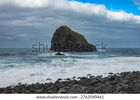 A dome sea stack rises from the Atlantic off Madeira north shore, dark basalt base with green top, foamy waves roll in, brooding clouds part near a bright horizon.