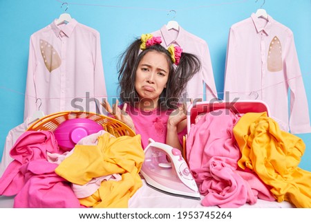 Domestic affairs. Displeased frustrated woman looks sadly at camera spreads palms brings large pile of laundry on ironing board feels tired of housework uses home appliance. Upset depressed housewife