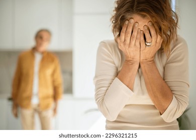 Domestic conflicts concept. Senior spouses quarreling in kitchen, elderly woman covering face with hands and crying while her husband standing on background, selective focus - Powered by Shutterstock