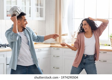 Domestic Fun. Cheerful arab couple playfully fighting in kitchen, using spatulas as a weapons, happy middle eastern spouses fooling together at home, enjoying time with each other, copy space