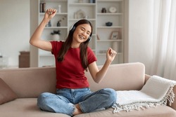 Domestic Fun. Cheerful Young Asian Woman Listening Music In Wireless Headphones While Relaxing On Couch At Home, Happy Korean Female Dancing And Singing While Resting On Sofa In Living Room