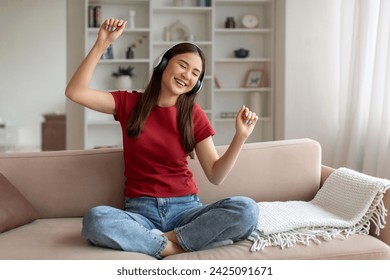 Domestic Fun. Cheerful Young Asian Woman Listening Music In Wireless Headphones While Relaxing On Couch At Home, Happy Korean Female Dancing And Singing While Resting On Sofa In Living Room - Powered by Shutterstock