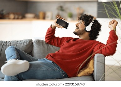 Domestic Fun. Happy Indian Man Listening Music In Wireless Headphones And Singing With Smartphone At Home, Cheerful Young Eastern Guy Relaxing On Couch In Living Room, Enjoying Favorite Playlist
