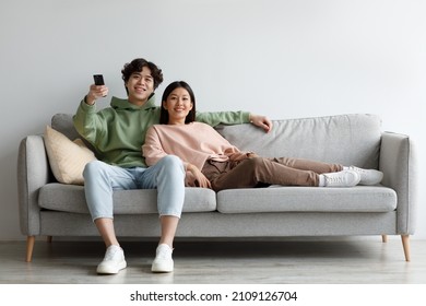 Domestic leisure activities. Positive young Asian couple relaxing on sofa, watching TV, switching channels with remote control, enjoying movie together at home, free space