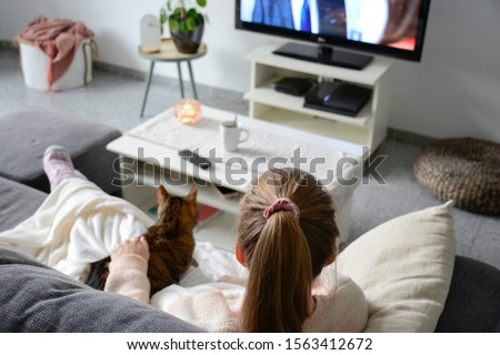Domestic life with pet at home. A young woman is sitting on the couch with her cat on her lap in the living room. She watches TV while stroking her cat. Binge watching tv via online streaming platform