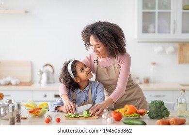 Domestic Life. Portrait of smiling African American woman helping daughter to prepare fresh healthy vegetable salad, little girl learning how to cook, chopping cucumber with knife on cutting board - Shutterstock ID 1945053862