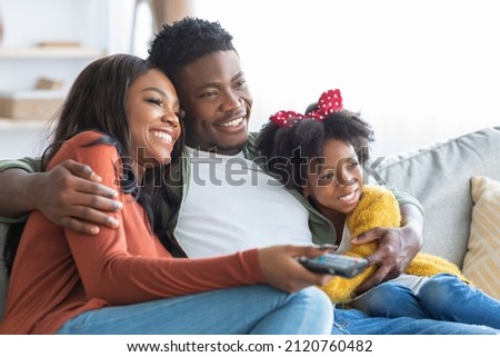 Domestic Pastime. Happy African American Family Of Three Watching Tv At Home, Young Black Parents And Cute Little Daughter Resting On Couch In Living Room, Enjoying Spending Time Together, Closeup