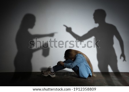 Domestic physical violence, abusing. Scared little caucasian girl, victim sitting close to white wall with shadow of angry threatening parents with alcohol addiction. Awareness of social problem.