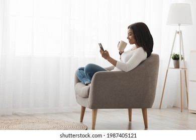 Domestic Rest. Calm Black Woman Using Smartphone, Sitting With Cup Of Coffee Near Window, Enjoying Morning, Free Space, Side View. Lady Surfing Internet Or Messaging Online On Hpone