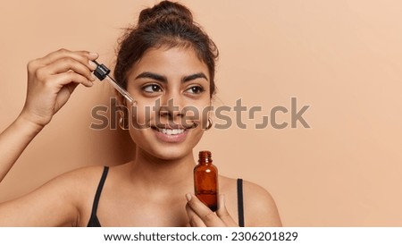 Domestic skin care and anti wrinkle routine. Pleased dark haired woman applies facial serum with dropper smiles toohthily focused aside dressed in t shirt isolated over brown background copy space