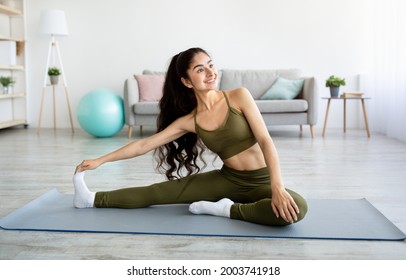 Domestic sports concept. Athletic Indian woman in sportswear doing fitness exercises on yoga mat, stretching her legs at home. Fit young Eastern lady staying flexible during covid lockdown