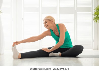 Domestic Sports Concept. Athletic senior woman in sportswear stretching on yoga mat, smiling mature female doing fitness exercises in living room, warming up leg muscles at home, free space - Powered by Shutterstock