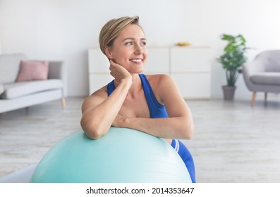 Domestic Train Concept. Smiling Woman Doing Exercises At Home, Leaning On Fitness Ball, Resting And Thinking, Looking Away. Caucasian Lady Working Out In Living Room With Sports Equipment