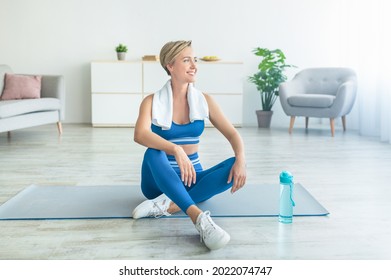 Domestic Train Concept. Smiling Woman Exercising At Home, Taking Break, Resting And Thinking, Looking Away At Window. Middle-aged Lady Sitting On Yoga Mat In Living Room With Towel On Shoulders