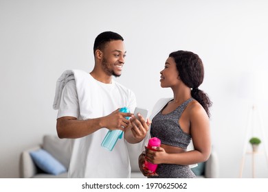 Domestic Training. Athletic Black Couple In Sportswear Holding Bottles Of Water, Using Smartphone, Ready For Workout At Home. Millennial African American Family Watching Sports Video On Mobile Device