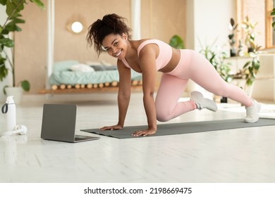 Domestic training concept. Happy fit black lady doing cross body mountain climbers exercise, watching online tutorial on laptop and smiling at camera, exercising at home - Powered by Shutterstock