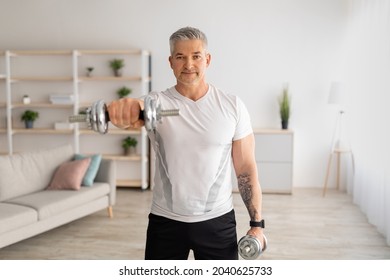 Domestic training with weights. Positive mature man doing exercises with dumbbells, strengthening his body at home. Senior sportsman working on his biceps muscles, staying healthy - Powered by Shutterstock