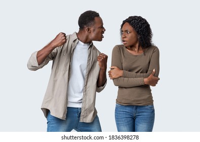 Domestic Violence And Abuse Concept. Young African American Mad Man Threatening His Woman With His Fist, Gray Studio Background. Furious Black Guy Showing Fists His Scared Girlfriend Or Wife