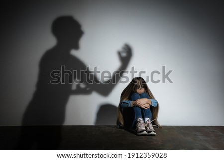 Domestic violence, abusing. Scared little caucasian girl, victim sitting close to white wall with shadow of angry father's threatening on it. Awareness of social problem, childhood, physical violence.