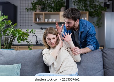 Domestic violence. Family conflict. A man shouts and beats a woman at home. Frightened woman closes her arms, sitting on the couch. She is afraid