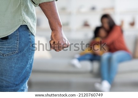Domestic Violence. Unrecognizable African American Man Threatening Wife And Daughter With His Fist, Scared Mother Embracing Little Girl While Sitting Together On Couch, Selective Focus On Male Hand