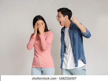 Domestic Violence. Young Asian Man Threatening His Girlfriend With His Fist, Gray Studio Background