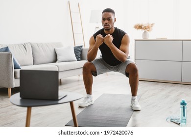 Domestic Workout. Athletic African American Guy Doing Deep Squats At Laptop During Online Training At Home. Male Fitness Routine. Sporty Black Man Exercising In Front Of Computer. Selective Focus