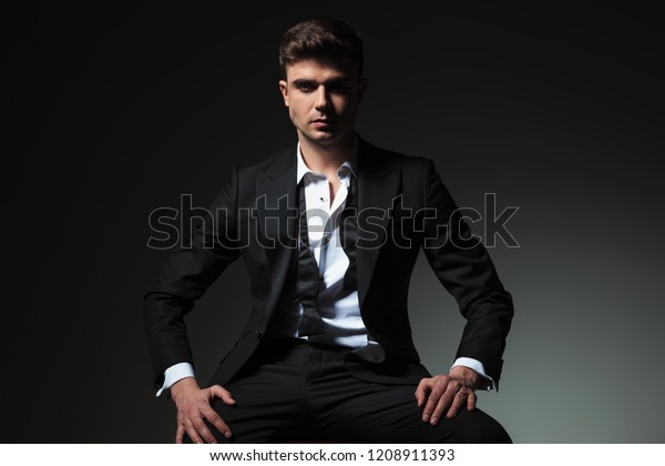 dominant man in\
black tuxedo with open collar and undone bowtie sitting on grey\
background with hands on\
thighs