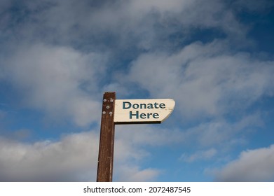 Donate Here Sign On White Painted Wood, On Dark Wood Post. Isolated Against Blue Sky And Clouds Background. Lots Of Copy Space.