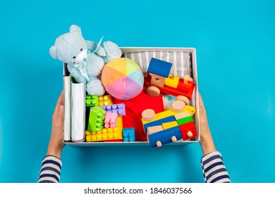 Donation Concept. Kid Hands With Donate Box With Clothes, Books And Toys On Light Blue Background. Top View